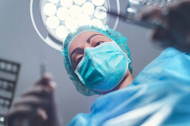Von unten Frau Chirurgin in medizinischen Uniform mit professionellen Werkzeugen, während stehend unter hellem Licht im Operationssaal - ADSF08418