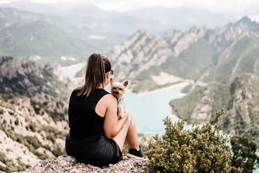 Rückenansicht eines anonymen weiblichen Touristen in lässiger schwarzer Kleidung und Sonnenbrille, der mit seinem Hund auf einem felsigen Hügel sitzt und die malerische Landschaft im Sommer bewundert - ADSF08444