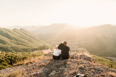 Back view of romantic couple of tourists in casual clothes sitting on stone edge of cliff embracing and enjoying picturesque landscape during sunny day - ADSF08445