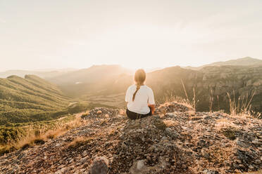 Back view of anonymous female tourist in casual clothes enjoying picturesque landscape while sitting on stony ground on sunny day in summer - ADSF08447