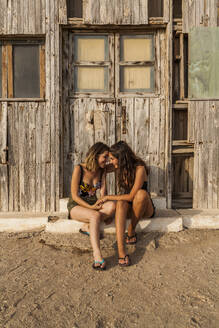 Zärtliches weibliches Paar, das an einem alten Holzschuppen sitzt, die Hände hält, sich umarmt, die Stirn berührt, miteinander spricht und lächelt, an einem sonnigen Tag, und sich gegenseitig ansieht - ADSF08462