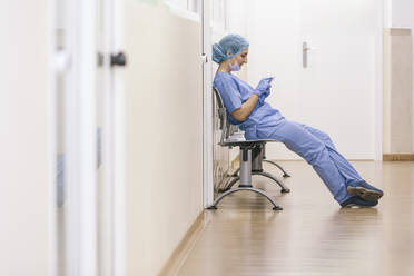 Female surgeon rests in a chair while checking messages on her smartphone - ADSF08501