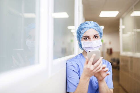 Female surgeon standing in the hallway while checking messages on her smart phone, look at camera and smile - ADSF08507