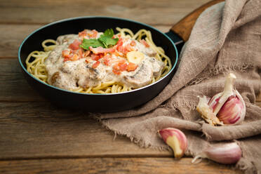 Von oben schmackhafte Spaghetti mit Schinkenscheiben und Champignons in cremiger Soße in der Pfanne gekocht und auf einem Holzbrett auf einem Holztisch mit Knoblauch und Leinenstoff zur Seite gestellt - ADSF08513