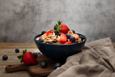 Von oben köstliches Frühstück Schüssel Cornflakes mit Erdbeeren und Blaubeeren auf Schneidebrett platziert und dekoriert mit Leinentuch und Beeren um Schüssel auf Holztisch mit grauem Hintergrund - ADSF08515