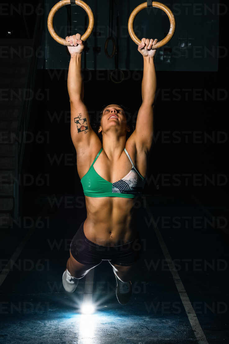 Full body strong sportswoman hanging on gymnastic rings and swinging while  exercising during intense training in dark gym stock photo