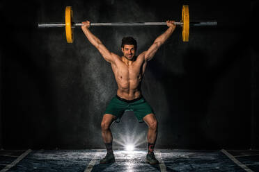Ein muskulöser männlicher Ganzkörpersportler lächelt und hebt eine schwere Langhantel während eines intensiven Krafttrainings in einem dunklen Fitnessstudio - ADSF08537
