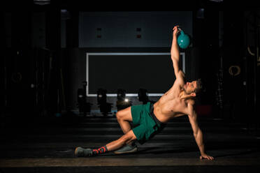 Muscular male athlete in shorts doing side plank and lifting kettlebell during functional workout in dark gym - ADSF08538