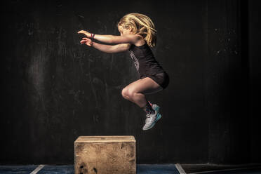Seitenansicht einer starken Frau in Sportkleidung, die während eines intensiven Trainings im Fitnessstudio auf einen schäbigen Holzkasten springt - ADSF08543
