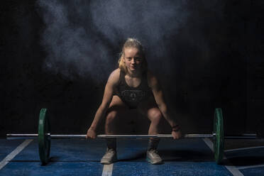 Glückliche muskulöse Frau gewinnt lächelnd Sportkleidung und hebt eine schwere Hantel über den Kopf während eines intensiven Trainings im dunklen Fitnessstudio - ADSF08544