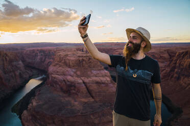 Handsome bearded man in hat cheerfully smiling and taking selfie while standing against magnificent canyon and river during sunset on West Coast of USA - ADSF08635