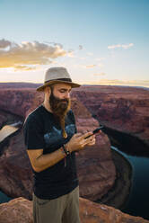 Handsome bearded man in hat on the mobile phone while standing against magnificent canyon and river during sunset on West Coast of USA - ADSF08636
