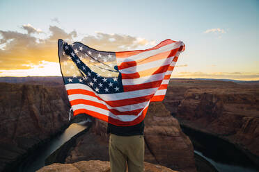 Back view of male with waving USA flag standing near beautiful canyon against sunset sky on West Coast - ADSF08639
