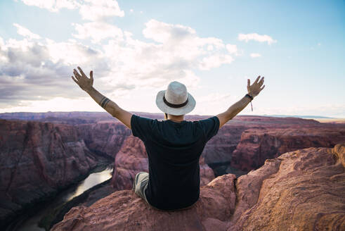 Rückenansicht eines Mannes mit Hut, der die Arme ausstreckt und die Aussicht auf einen majestätischen Canyon gegen den wolkenverhangenen blauen Himmel bewundert, während er durch die Westküste der USA reist - ADSF08644