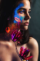 Scary young female in colorful contact lenses and with painted face  standing in dark forest and