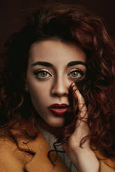 Young curly brown haired female in casual holding strand of hair in hand next to face while looking at camera with big grey eyes - ADSF08750