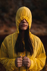 Female in yellow rain coat wearing hood covering face standing in nature on cloudy day - ADSF08753