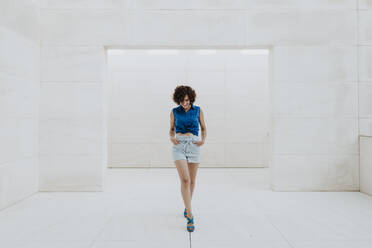 Stylish woman with curly hair walking on tiled floor against wall - GMLF00378