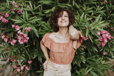 Close-up of smiling mid adult woman with curly hair standing against plants in park - GMLF00390