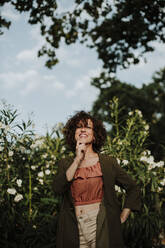 Lächelnde mittelgroße erwachsene Frau mit lockigem Haar und Mantel, die gegen Pflanzen im Park steht - GMLF00395