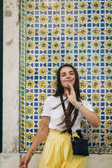 Lächelnde schöne Frau mit langen Haaren, die an einer gekachelten Wand in einer Stadt steht - DCRF00570