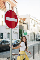 Cheerful beautiful woman with bicycle standing by road sign in city - DCRF00572