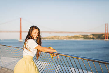 Lächelnde schöne Frau mit langen Haaren steht am Geländer über den Fluss in der Stadt bei Sonnenuntergang - DCRF00576