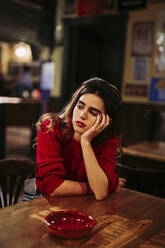 Sad thoughtful mid adult woman with hand on chin sitting at table in restaurant - LJF01717