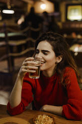 Nahaufnahme einer lächelnden Frau, die ein Bierglas in der Hand hält, während sie an einem Tisch im Restaurant sitzt - LJF01724