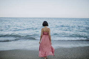Frau im Kleid geht am Strand gegen den klaren Himmel bei Sonnenuntergang zum Meer - GMLF00402