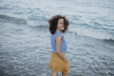 Fröhliche mittelgroße erwachsene Frau mit lockigem Haar im Meer stehend bei Sonnenuntergang - GMLF00404