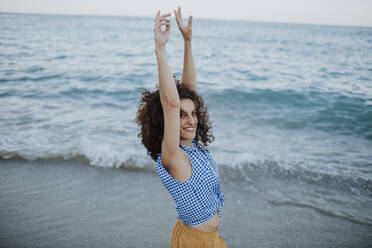 Smiling mid adult woman with arms raised standing against sea during sunset - GMLF00405