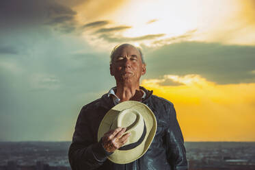 Senior man holding hat against cloudy sky during sunset - OCMF01595