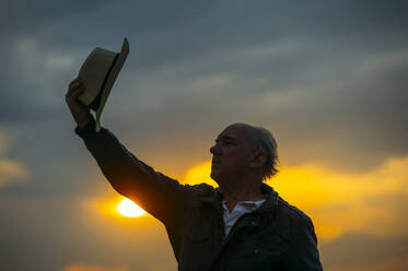 Älterer Mann hebt seinen Hut gegen den Himmel bei Sonnenuntergang - OCMF01597