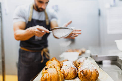 Baker spraying powdered sugar on croissants at bakery - MRRF00190