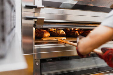 Abgeschnittene Hand eines männlichen Bäckers, der in einer Bäckerei Brot im Ofen backt - MRRF00191