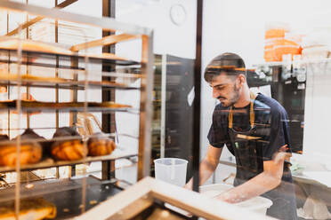 Junger Bäcker bei der Arbeit in einer Bäckerei durch ein Glasfenster gesehen - MRRF00198