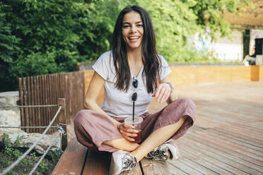 Junge fröhliche Frau mit Kaffee in der Hand auf einer Bank im Park sitzend - OYF00179