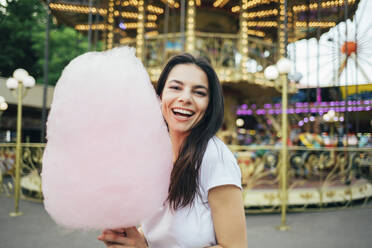 Cheerful beautiful woman holding cotton candy standing against carousel in amusement park - OYF00182