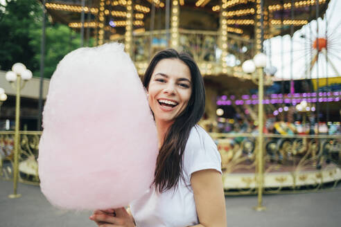 Fröhliche schöne Frau mit Zuckerwatte vor einem Karussell in einem Vergnügungspark - OYF00182