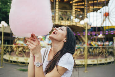 Happy young woman holding cotton candy standing in amusement park - OYF00183