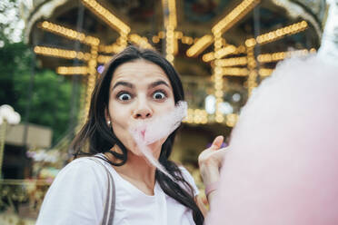 Close-up of young woman eating cotton candy against carousel in amusement park - OYF00187