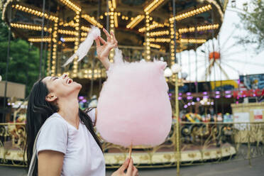 Happy beautiful woman eating cotton candy while standing against carousel in amusement park - OYF00188
