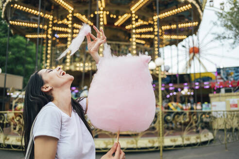 Glückliche schöne Frau, die Zuckerwatte isst, während sie vor einem Karussell im Vergnügungspark steht - OYF00188