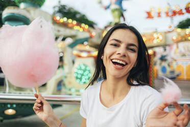 Nahaufnahme einer schönen Frau, die lachend Zuckerwatte in einem Vergnügungspark hält - OYF00190