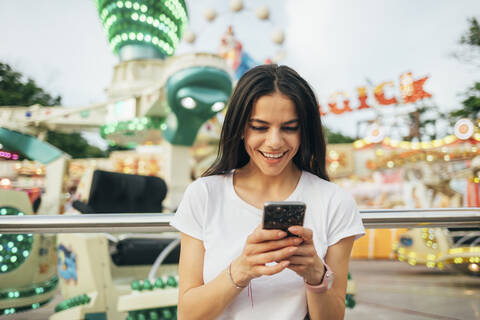 Lächelnde junge Frau, die ein Smartphone benutzt, während sie in einem Vergnügungspark steht, lizenzfreies Stockfoto