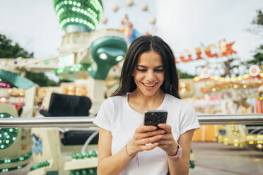 Lächelnde junge Frau, die ein Smartphone benutzt, während sie in einem Vergnügungspark steht - OYF00193