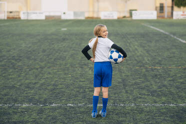 Mädchen in weißer und blauer Uniform mit Fußball lächelt in die Kamera, während sie allein auf einem grünen Feld in einem modernen Sportverein steht - ADSF08781