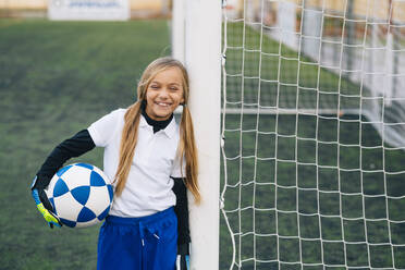 Fröhliches Mädchen in weißer und blauer Uniform mit Fußball, das in die Kamera lächelt, während es allein auf einem grünen Feld in einem modernen Sportverein steht - ADSF08782