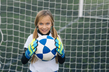 Fröhliches Mädchen in weißer und blauer Uniform mit Fußball, das in die Kamera lächelt, während es allein auf einem grünen Feld in einem modernen Sportverein steht - ADSF08783